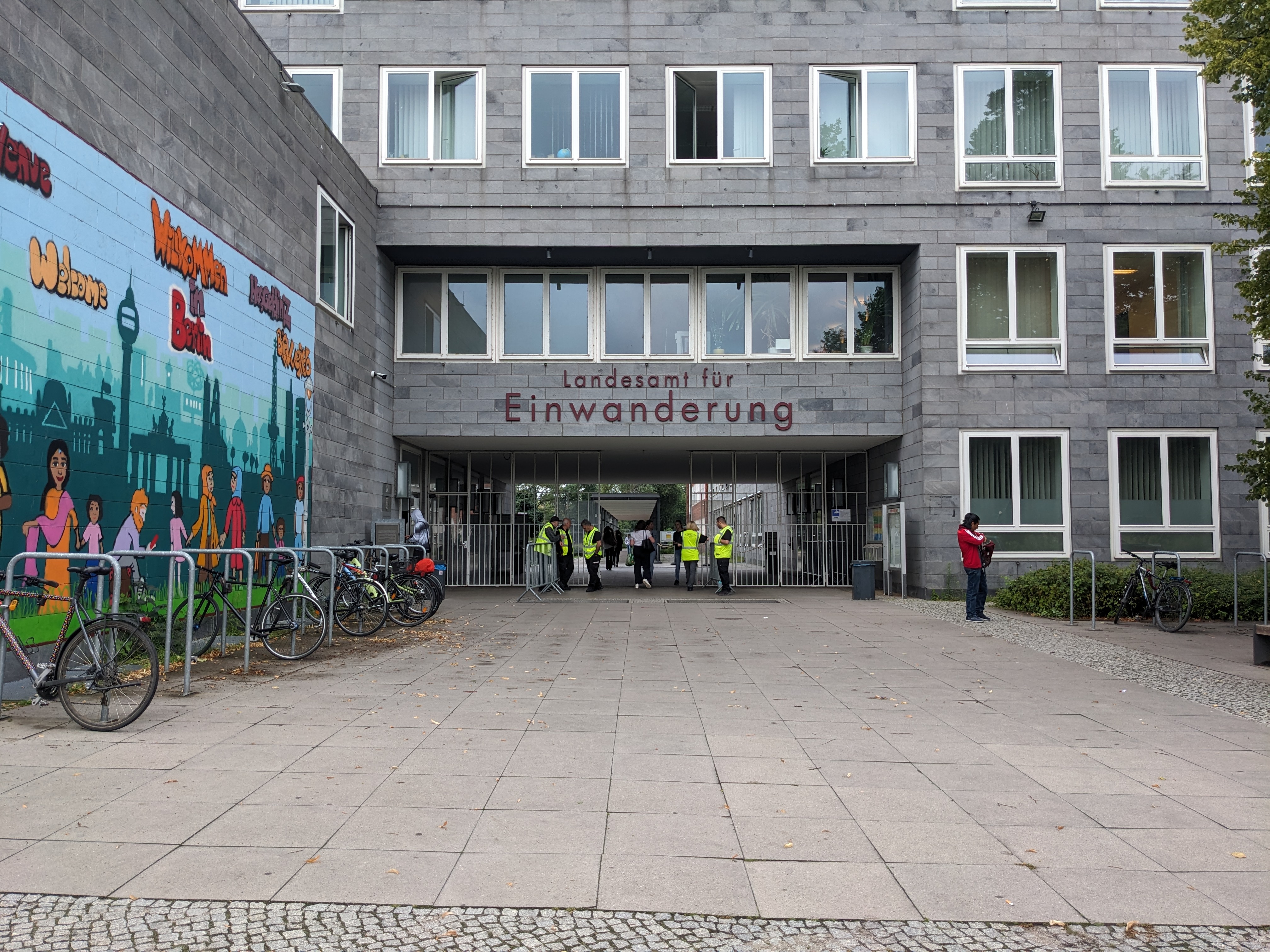 مدخل مكتب الهجرة الألماني (Landesamt für Einwanderung) مع لوحة جدارية ملونة على الجدار الأيسر ودراجات متوقفة بالخارج.